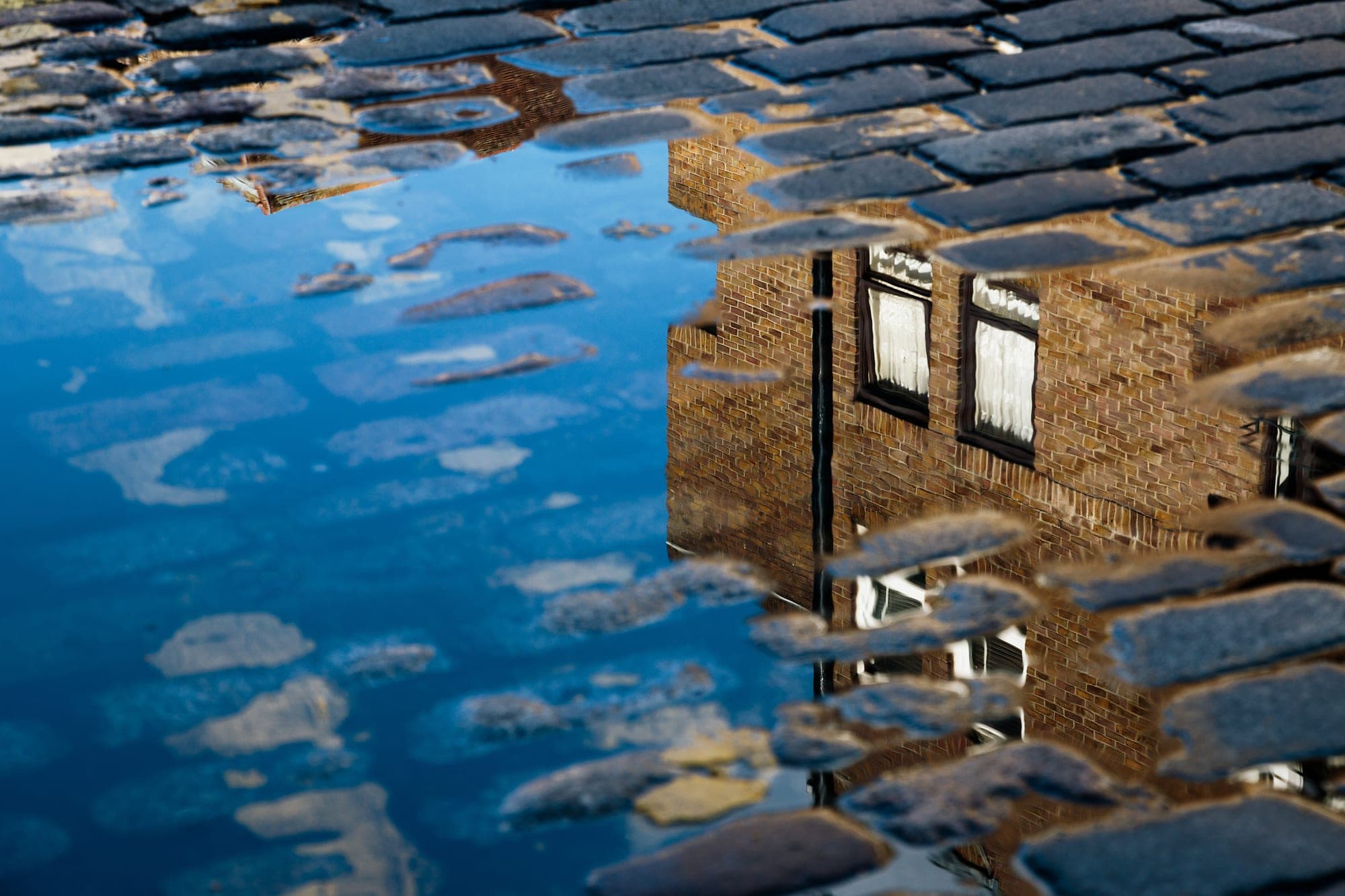 A brick building reflected in a puddle between cobblestones