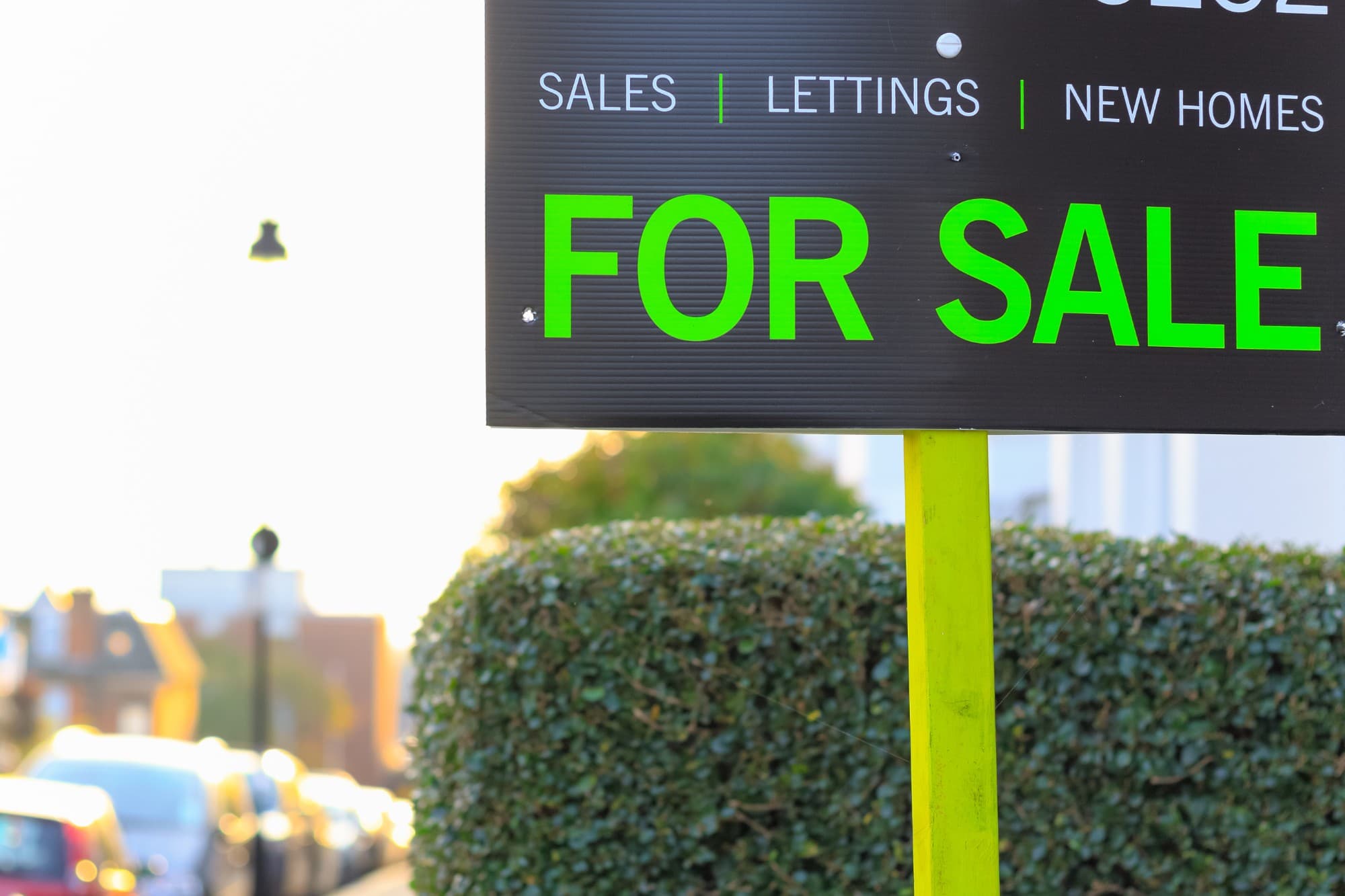An estate agent's For Sale board on a post beside a hedge on a residential street.