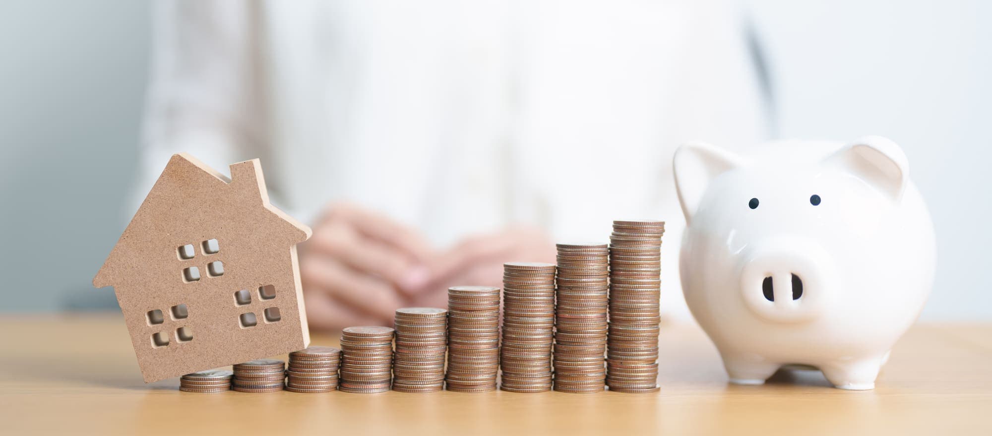 A wooden house beside rising stacks of coins and a piggy bank